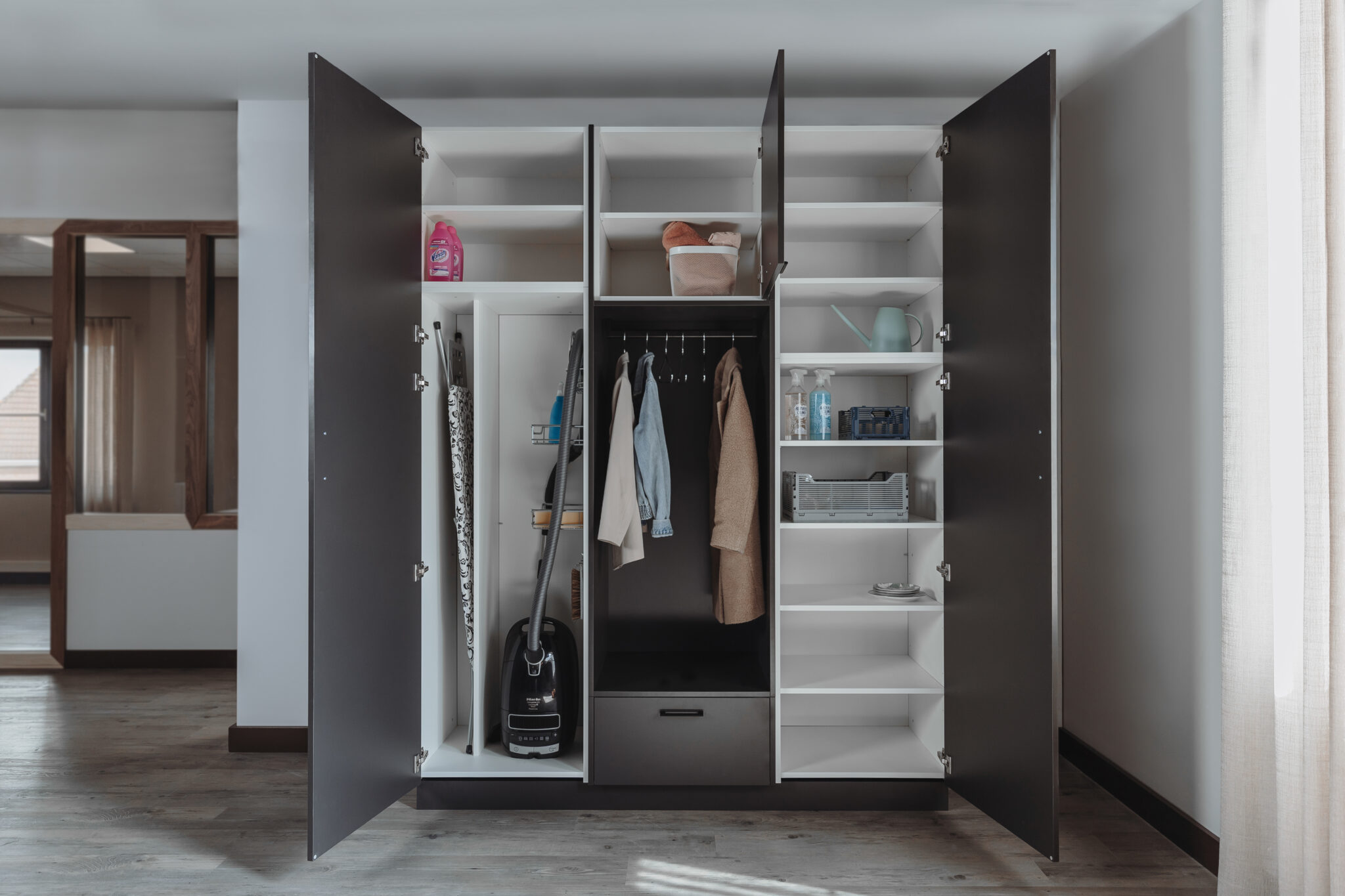 Bijkeukenkast met open deuren, gevuld met schoonmaakmiddelen, kleding en huishoudelijke spullen.