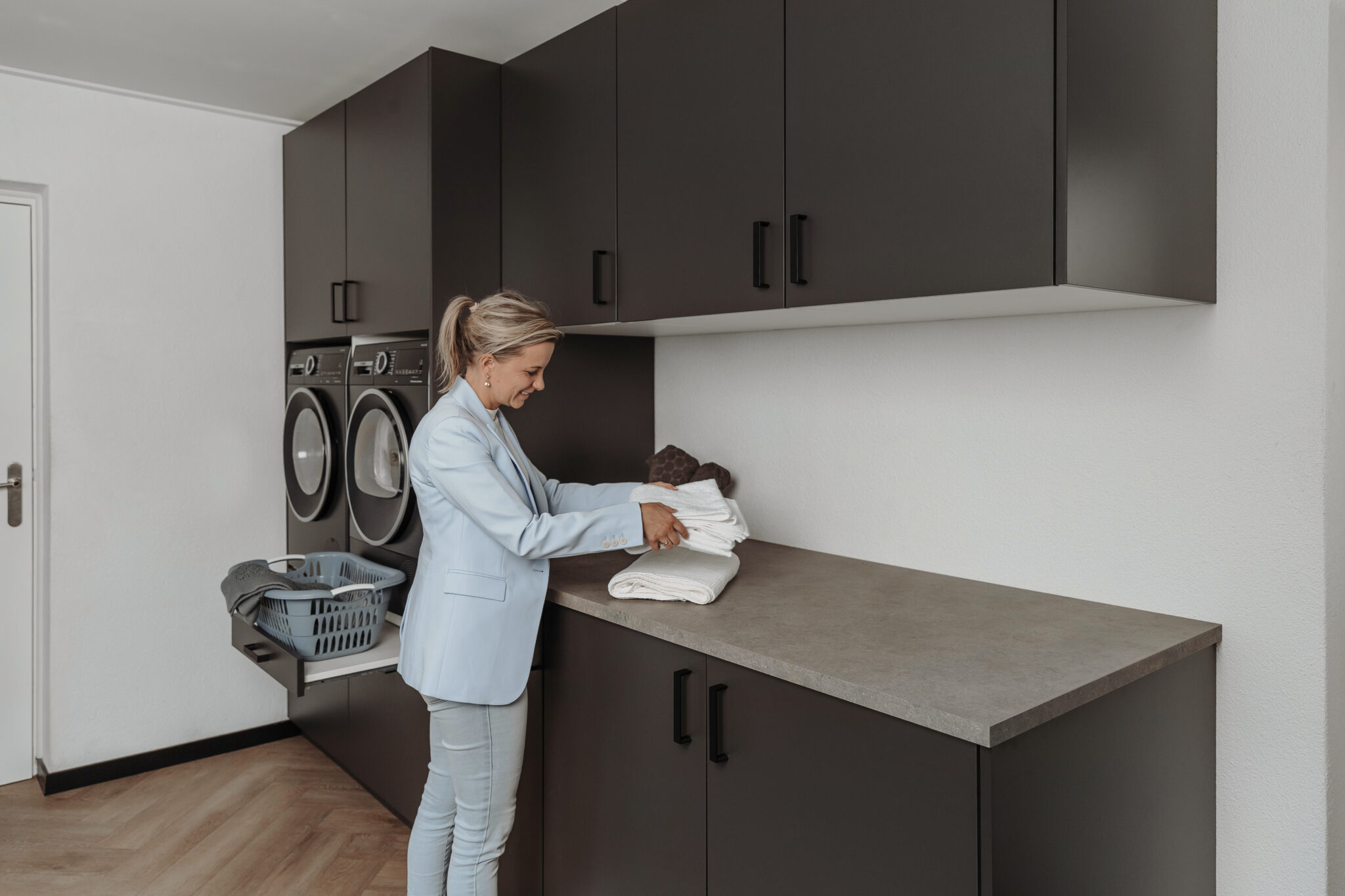 Lachende vrouw in lichtblauw colbert vouwt schone witte handdoeken op het aanrecht van een moderne wasruimte met donkergrijze kasten, ingebouwde wasmachine en droger, en een uitschuifbare plank met wasmand.